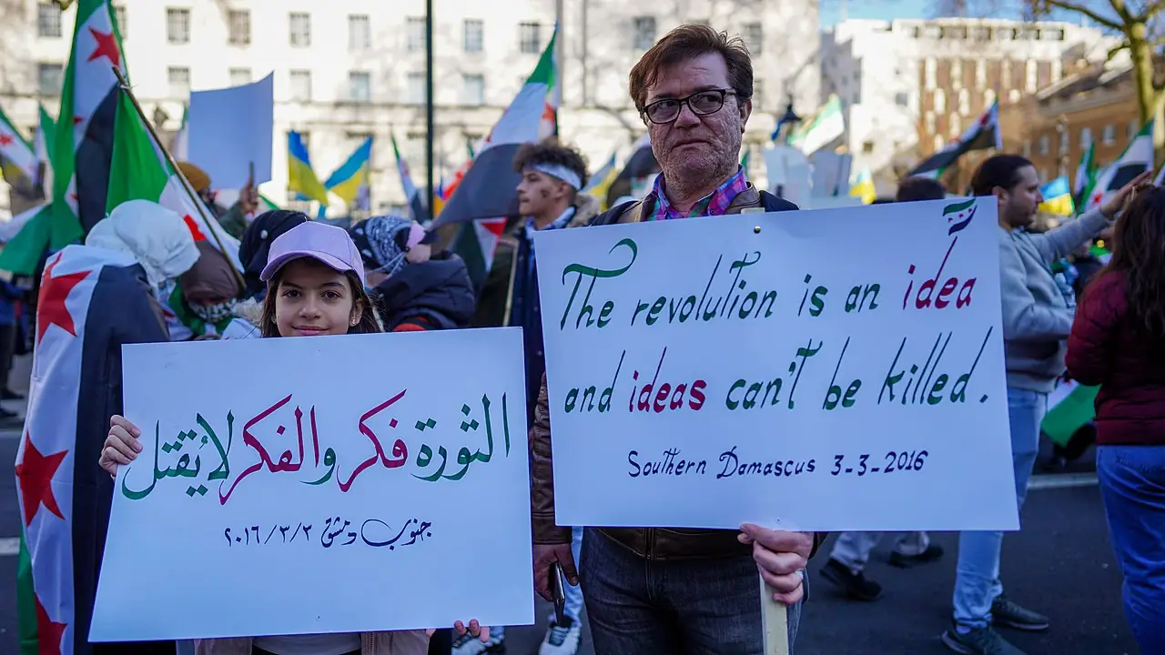 Protesters on the streets wielding signs. A child and an adult, with the adult showcasing a quote reading 'The revolution is an idea and ideas can't be killed.' 