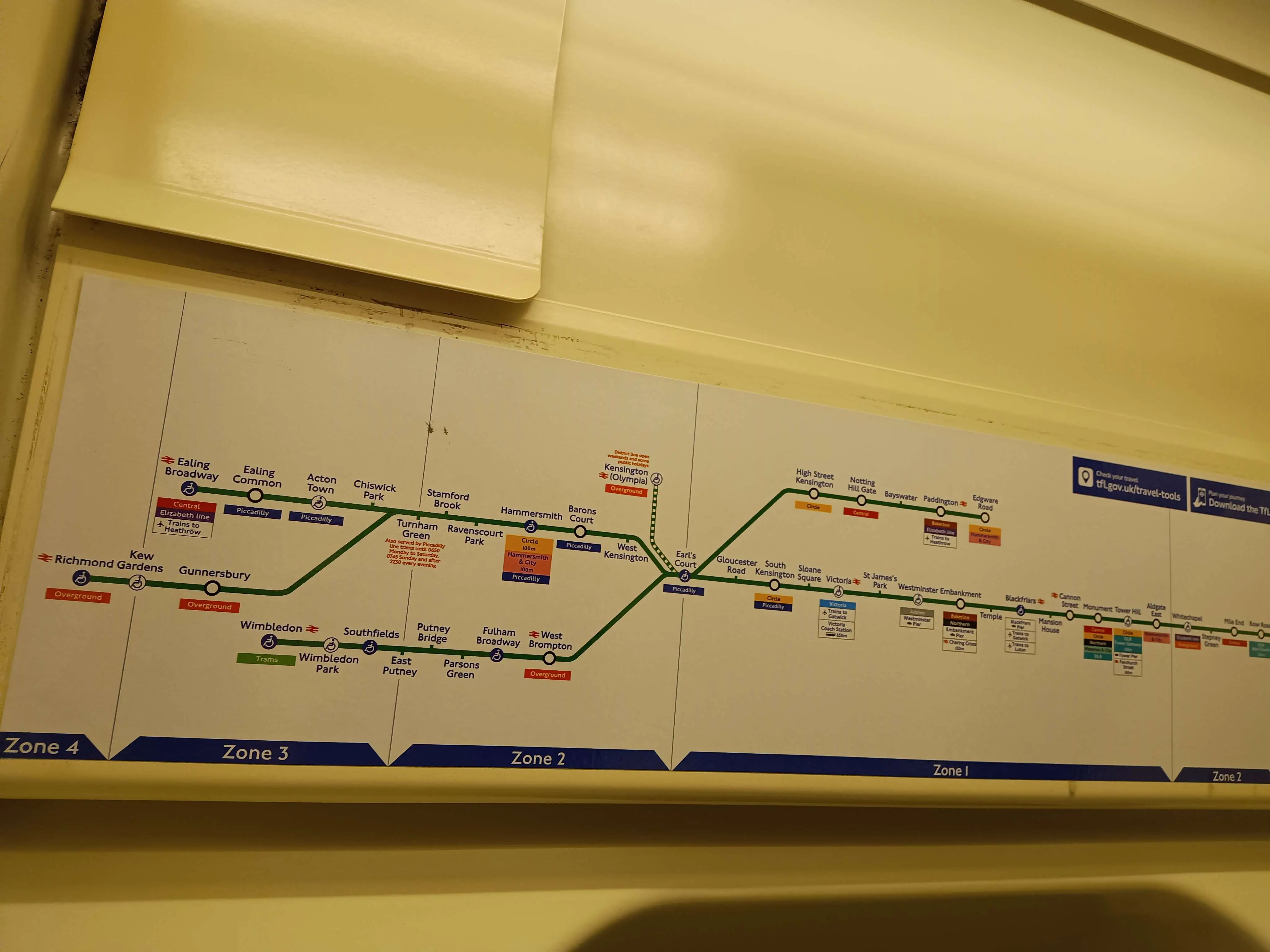 A District line map displayed against a beige looking tube wall, most of the line is coloured solid green while the branch, going to the Kensington Olympia stop, is displayed with white edges in between the green to mark it out, plus red text reading: District line open weekends and some public holidays.